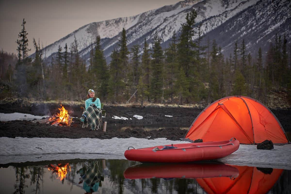 View of camp life. Woman sitting near campfire, drinking tea or coffe and have a rest. Orange tent next to lake shore. Canoe on water near the tent. Snow mountains on background.