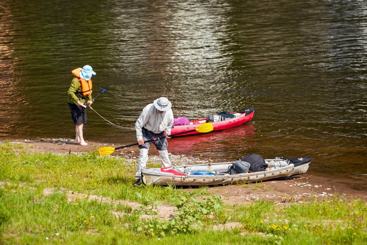 Two kayakers take a rest on the banks of a river. They are well equipped with life jackets, hats, luggage and a red and white kaja or canoe