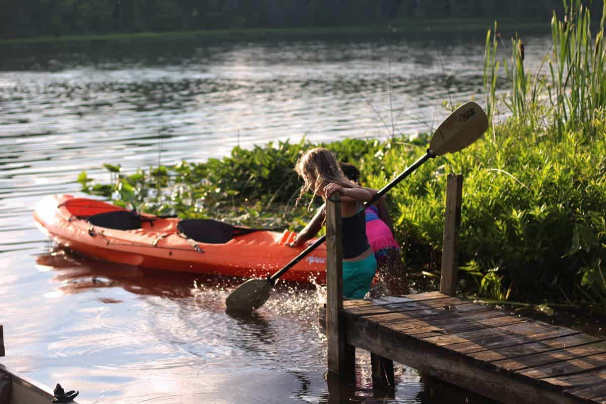 Two children wading to water towards orange kayak with black kayak seat