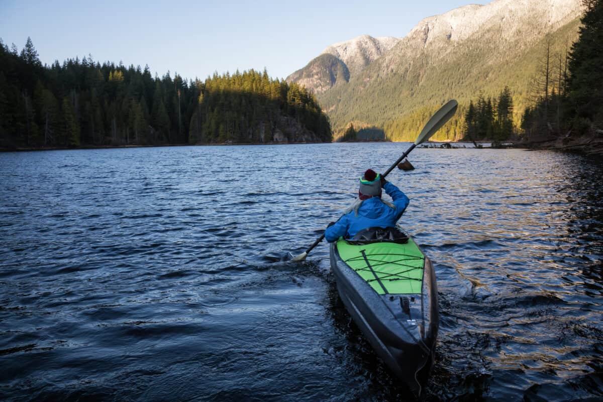 Lady in bobble hat paddles a rigid frame inflatable kayak on a lake surrounded by mountains