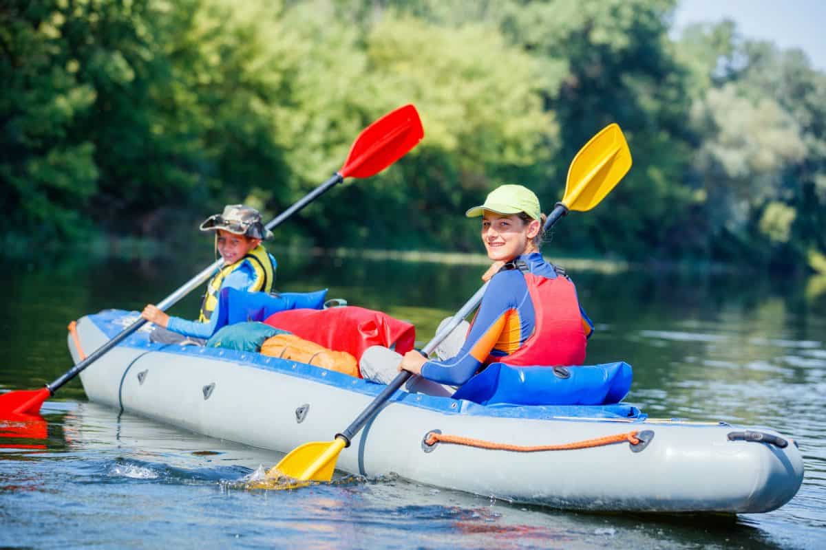 Two children both who are wearing a life jacket paddle a kayak on the river
